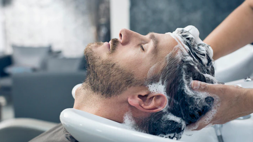 A bearded man with his eyes closed and his head covered in shampoo lather reclines at a salon sink, as a stylist's hands massage his hair.