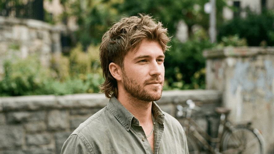 A man outdoors modeling a modern mullet hairstyle, featuring a textured, slightly messy top with shorter sides and a softly blended longer back.